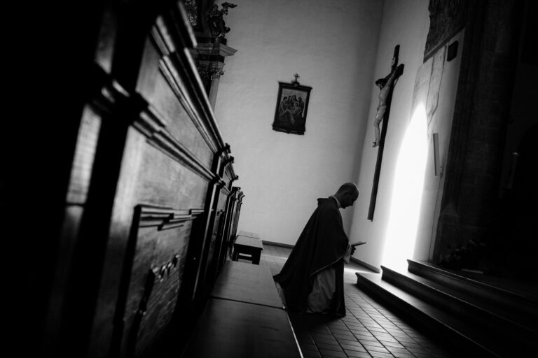 Náhled do interiéru starého kostela s klasicistním oltářem a sochou Krista. Lidé jsou vidět v pozadí, při modlitbě nebo jiné formě bohoslužby. Umělecké detaily vytváří kontrast mezi světlem a stínem.