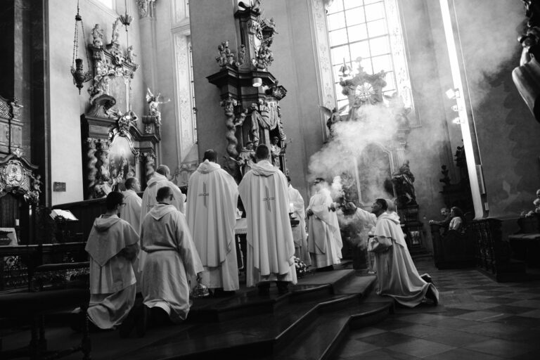 Pohled do interiéru farního kostela s ministranty a duchovními při bohoslužbě. Kouř kadidla se vznáší prostorem kostela.