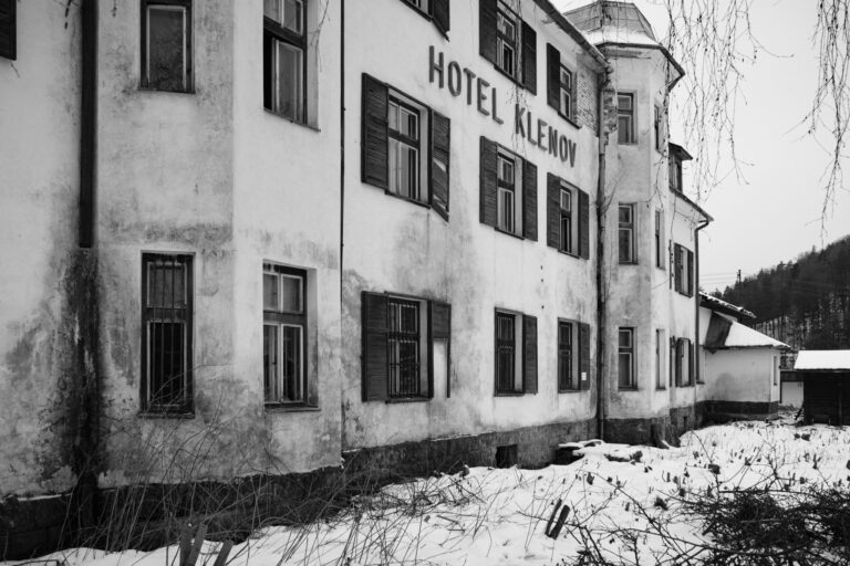 Pohled na bílý hotel s nápisy HOTEL KLENOV. Budova má několik podlaží. Nachází se v zimním prostředí s sněhem na zemi. Obrysy okolních budov a stromů jsou viditelné na pozadí.
