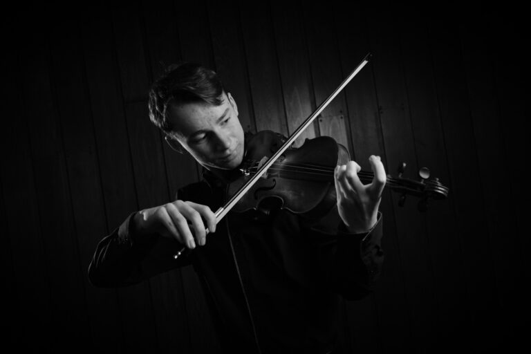 Muž hrající housle v černobílém snímku. Houslista se soustředěně hledí na nástroj a ruce drží housle a hudební skladbu.
