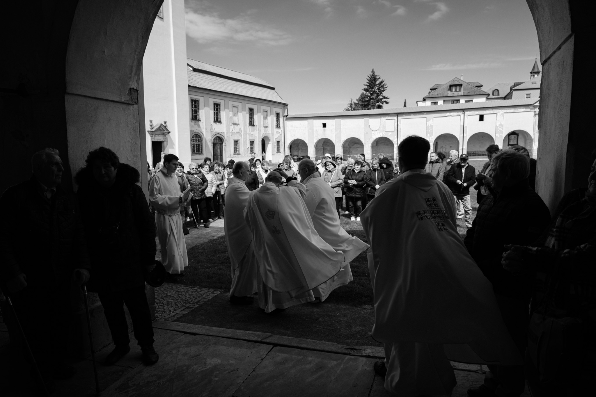 Fotografie zachycuje skupinu lidí před kostelem. Viditelné jsou postavy v liturgických oděvech. V popředí je část budovy kostela s dveřmi a okny. V pozadí je vidět krajina. Na fotografii jsou lidé, kostel, priest, Mons. František Rábek a Sv. Kopeček.